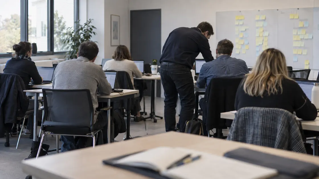 Atelier de sensibilisation : des participants travaillent sur leurs ordinateurs portables pendant que le formateur, debout, accompagne l’un d’eux. Au mur, des post-it de tâches métier.